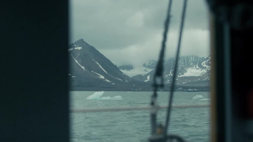 Peeking through a ship's window, a slow pan reveals a vast arctic landscape with snow-capped mountains and floating icebergs on a cloudy day