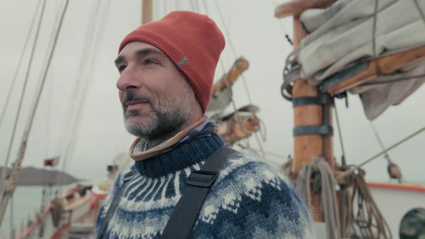 Portrait of a seasoned sailor in a warm sweater and beanie, looking thoughtfully from the deck of his wooden boat on an overcast day