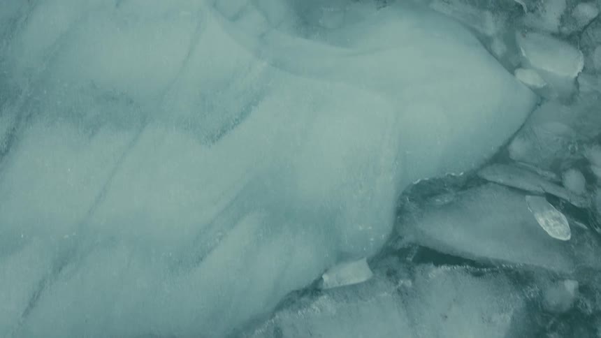An abstract top-down shot slowly pans over the cracked and layered turquoise ice of a frozen body of water