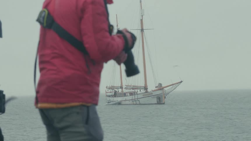 A photographer in a red jacket takes pictures of a large sailboat on the water during a foggy day