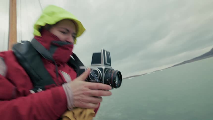 An adventurous photographer in a red jacket uses a classic film camera to capture the rugged arctic scenery from a sailboat