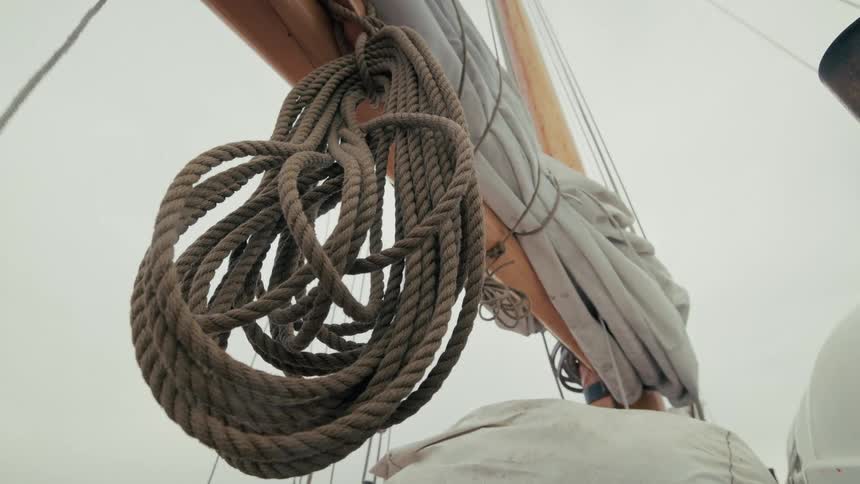 A coiled rope hangs from the boom of a sailboat next to a furled sail against an overcast sky