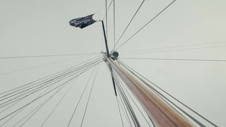 A black flag waves in the wind atop the tall wooden mast of a historic sailing ship against an overcast sky