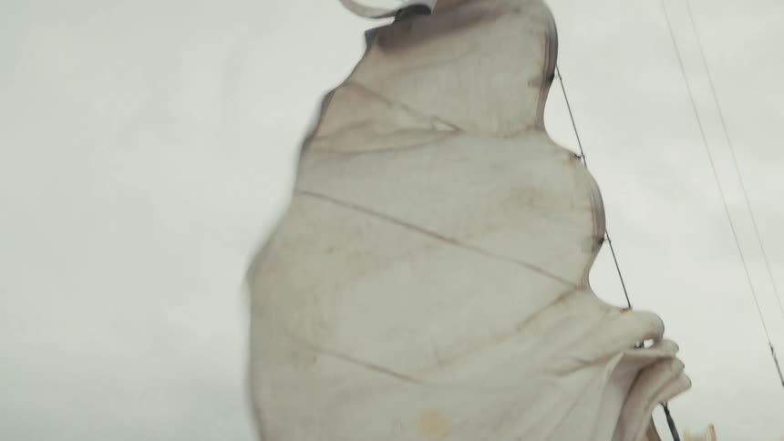 A large, weathered sail flaps wildly in the wind on a sailboat crossing the ocean on a gray, overcast day