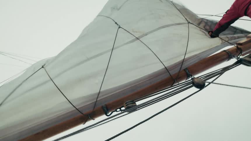 A sailor in a red survival suit works to furl the mainsail of a traditional wooden ship against an overcast sky