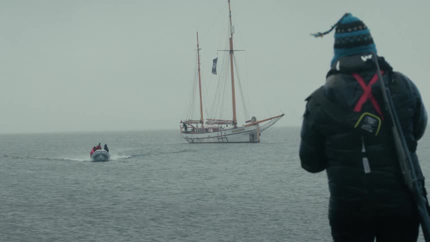A hunter with a rifle on their back watches from the shore as a small boat travels towards a large sailboat on the open sea