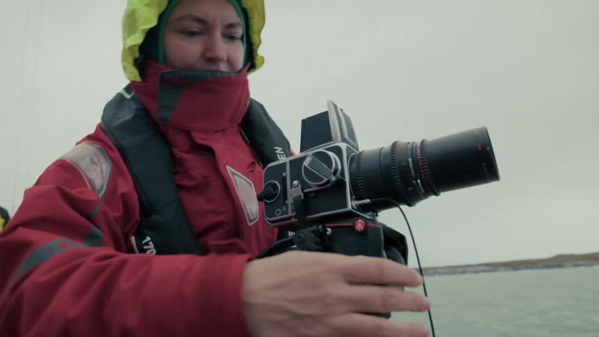 A professional photographer adjusts her medium-format film camera on a tripod while sailing in a cold, remote location