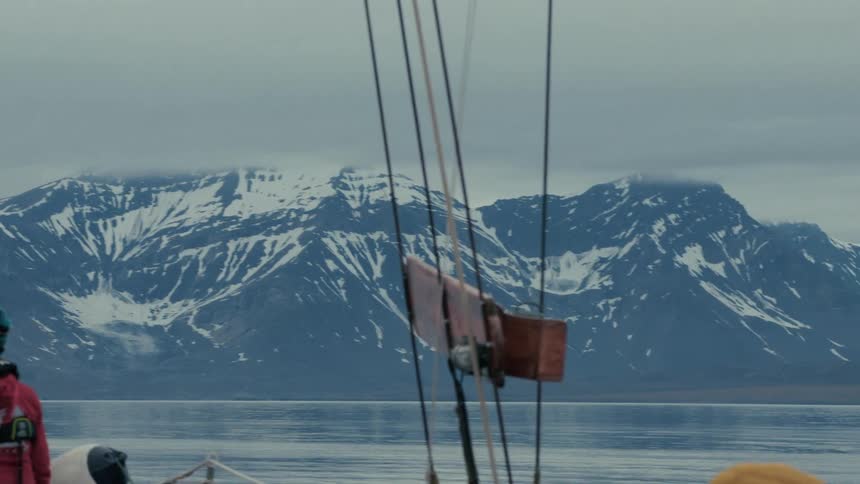 A person on a sailboat admires the vast, snow-covered mountains of the Arctic on a calm, overcast day