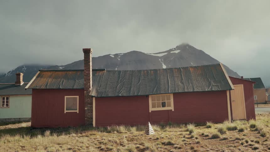 A quiet red cabin with a brick chimney sits in a desolate field with dramatic snow-capped mountains in the background