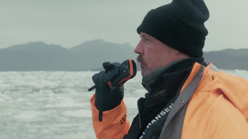 A man on a boat in an icy sea speaks into a walkie-talkie with mountains in the background