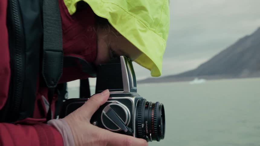 A photographer wearing a bright yellow hood uses a vintage film camera to capture the icy arctic landscape from a boat