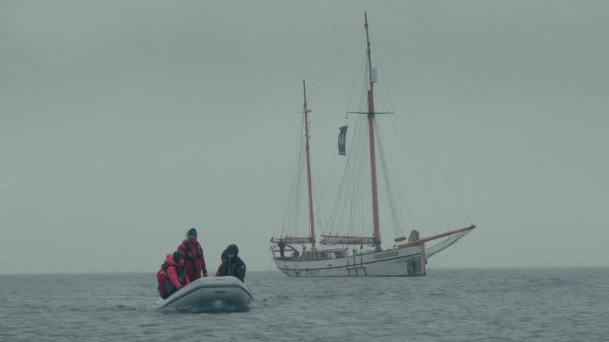 An expedition crew in a small inflatable boat floats on the ocean with a large schooner in the background on a foggy day