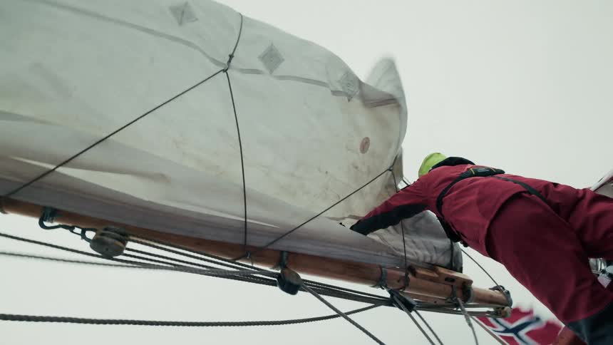 From a low angle, a sailor in a red suit lies on the boom to furl the flapping mainsail of a wooden sailboat on a foggy day