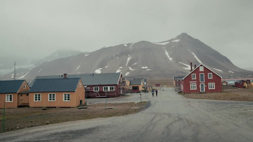Two people walk down the quiet street of a colorful, remote village with a large, snow-dusted mountain in the background