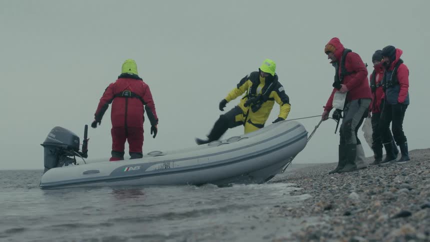 A team of explorers in heavy winter gear work together to bring their inflatable boat ashore on a pebble beach on an overcast day