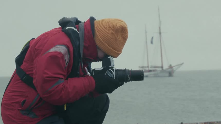 A photographer wearing a red jacket and yellow beanie crouches on a rocky shore to take a picture of a sailboat with a vintage camera