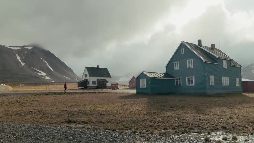 A solitary figure in a red jacket walks through a remote arctic village with colorful houses at the base of snowy mountains
