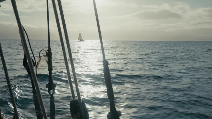 A distant sailboat glides across the glistening ocean at sunset, viewed through the rigging of another vessel