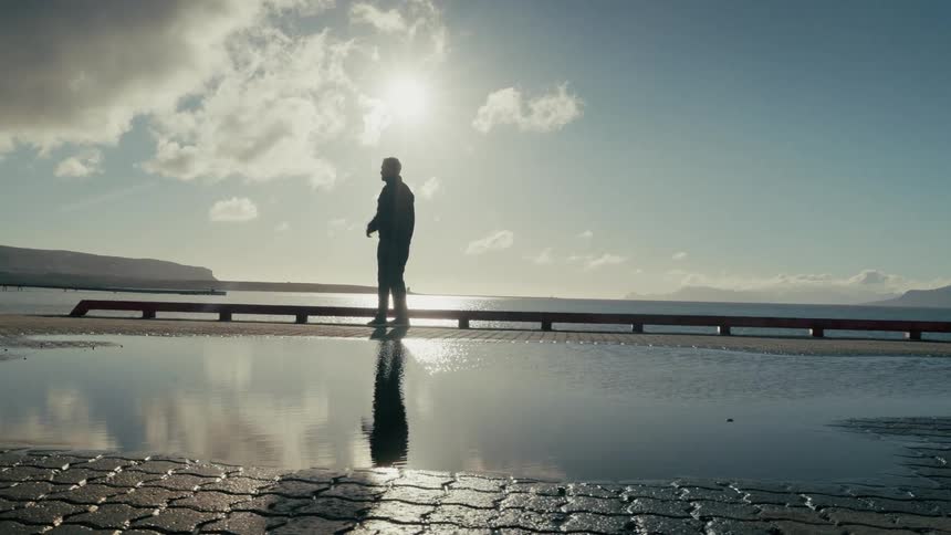 A man in silhouette stands on a pier looking out at the bright sun over the water, his reflection mirrored in a still puddle