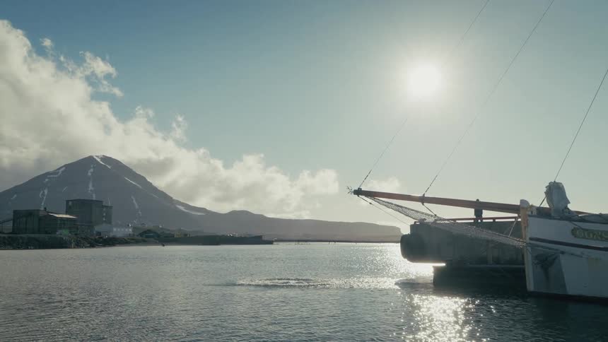 Slow motion footage shows an adventurous man jumping from the bow of a ship into the icy water of an arctic harbor with a mountain in the background