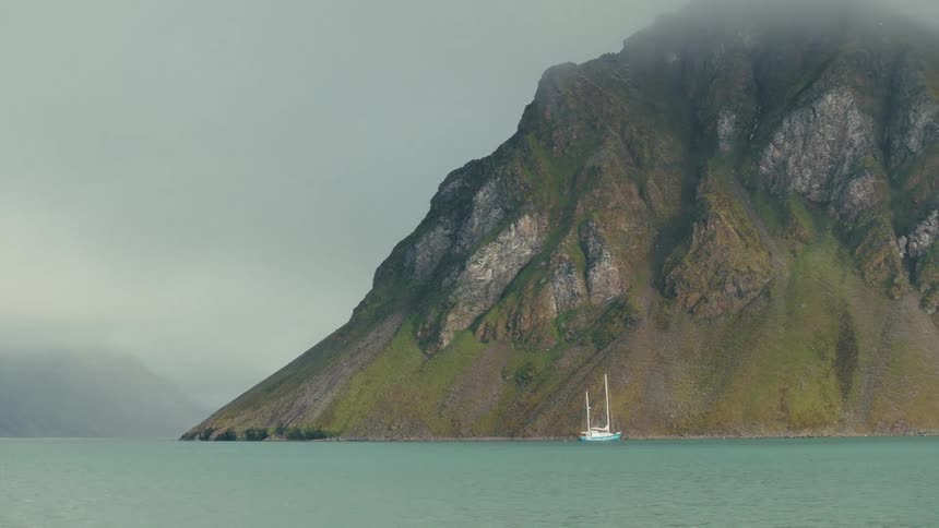 A sailboat is anchored in a serene fjord at the base of a massive, fog-shrouded green mountain