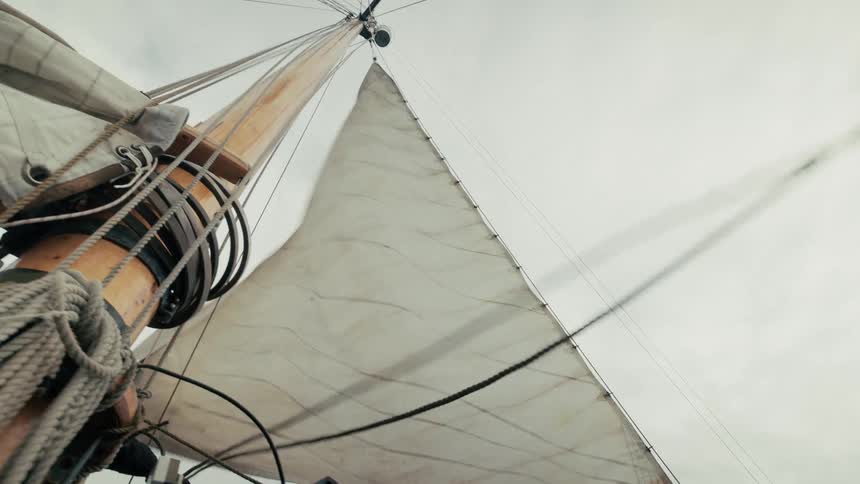 Looking up at the mast and main sail of a classic wooden boat as it sails on the water under an overcast sky