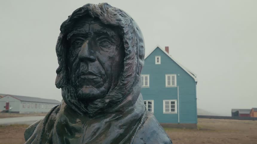 A stoic bronze bust of a hooded arctic explorer stands in a remote northern village on a foggy, overcast day