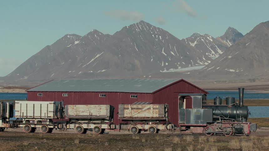 An old, abandoned steam train sits on display in front of a red barn with majestic, snow-capped mountains in the background