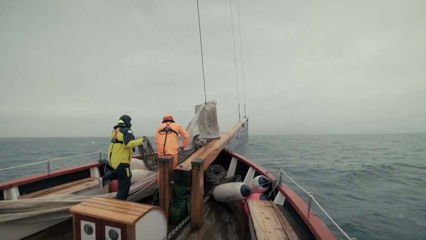 Two sailors in rain gear work together on the bow of a wooden boat as it sails across the open ocean on a cloudy day