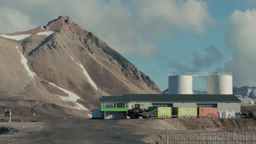 A remote industrial facility with a parked front loader and storage tanks operates at the base of a snowy mountain range