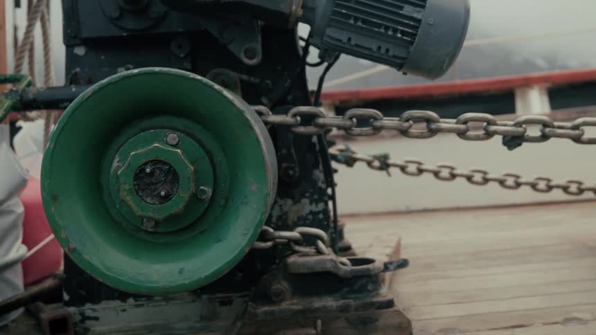 Close-up of a green anchor windlass slowly pulling a heavy chain on the wooden deck of a boat