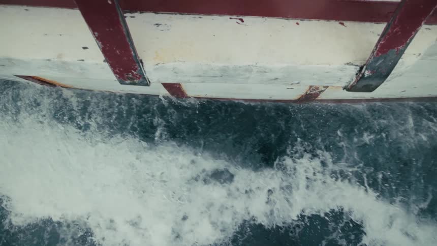 A top-down slow-motion shot of the side of a boat hull cutting through the dark ocean water, creating a churning wake