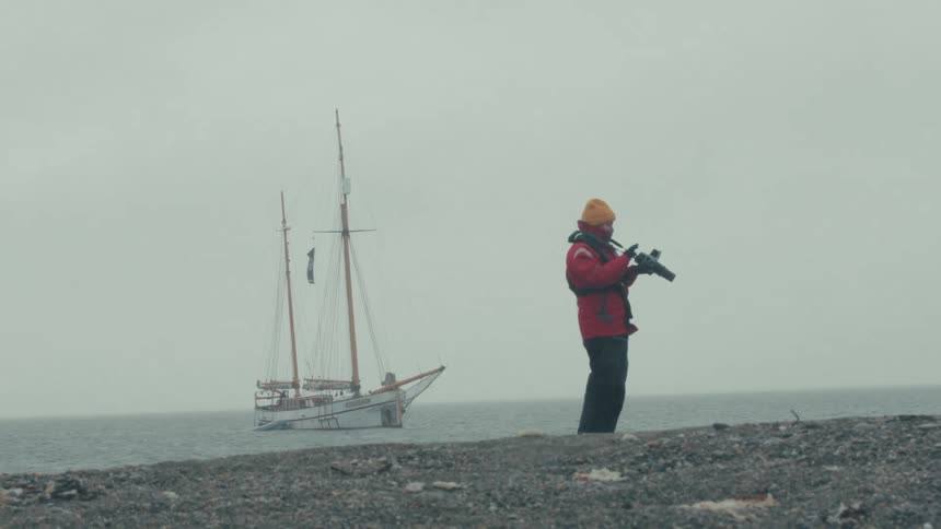 A polar bear guard stands watch with a rifle on a desolate arctic shore, with a two-masted sailboat anchored in the foggy sea behind