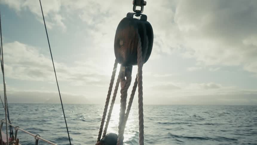 A close-up view of a wooden pulley on a sailboat with the sun shining brightly over the open ocean in the background