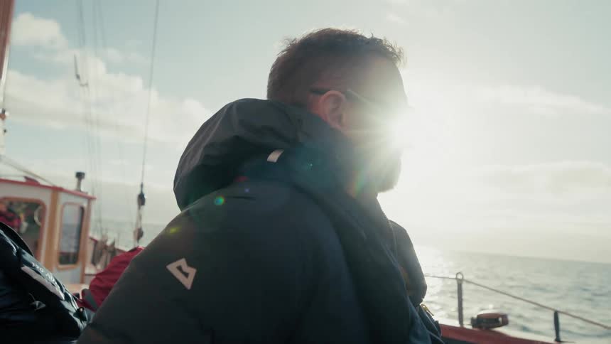 A man in a warm jacket and sunglasses enjoys the sunny view from the deck of a boat sailing on a calm ocean