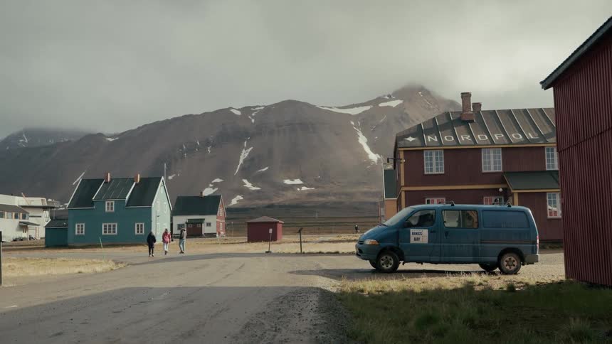 A quiet street in a remote Nordic village with colorful houses, a parked van, and people walking against a backdrop of snowy mountains