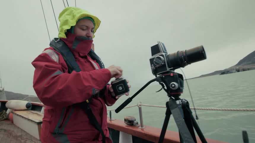 A photographer in a red rain jacket prepares her medium format film camera on the deck of a boat in an overcast sea