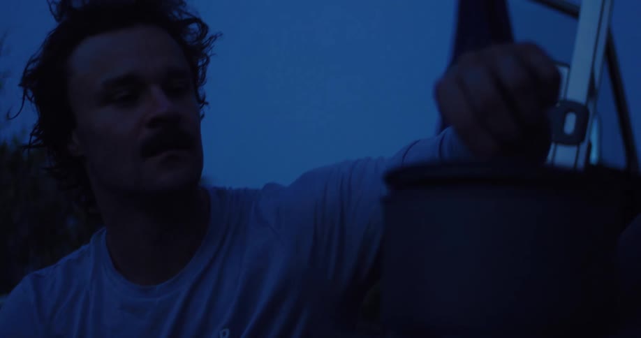 A weary cyclist with a mustache cooks his dinner on a portable stove at his campsite during the blue hour