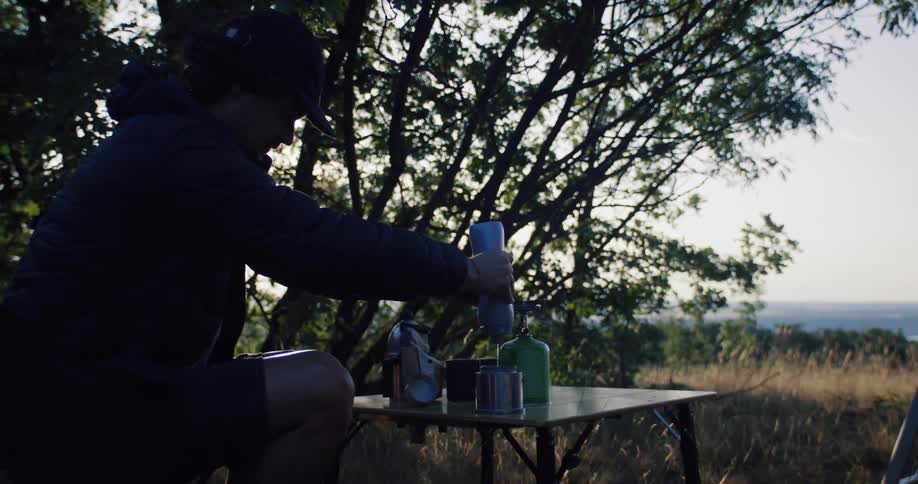 A silhouetted camper prepares his meal, pouring water into a pot on his camp stove as the sun sets