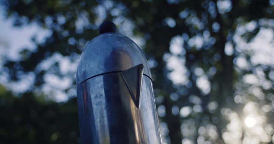 A silver moka pot lets off steam while brewing coffee outdoors with trees and sunlight in the background