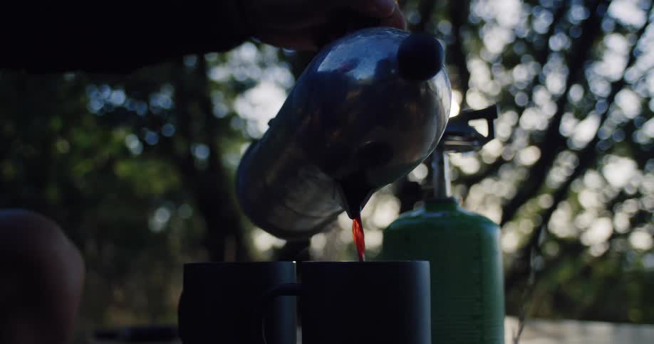 A person pours steaming hot coffee from a French press into a mug at a campsite in the early morning light