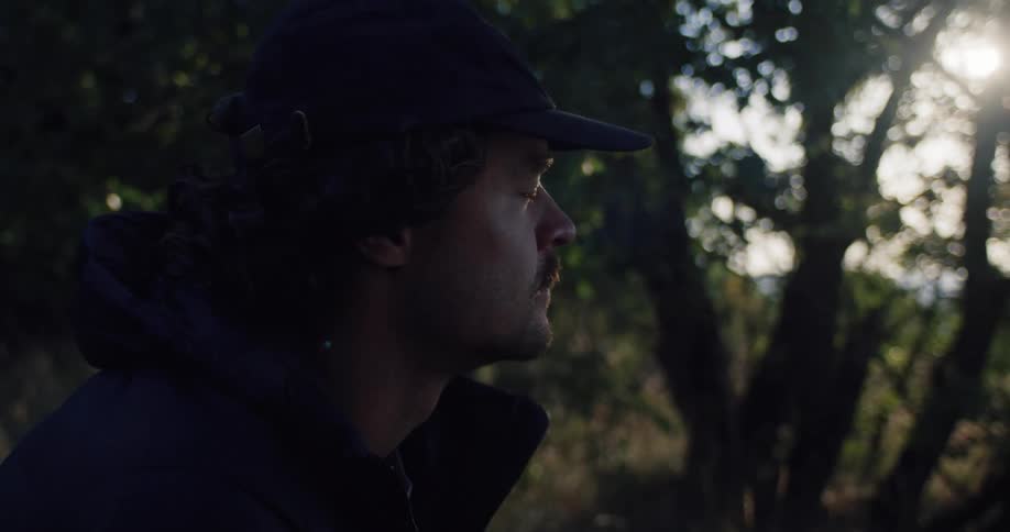 A thoughtful young man in a cap contemplates while standing in a quiet forest as the sun peeks through the trees