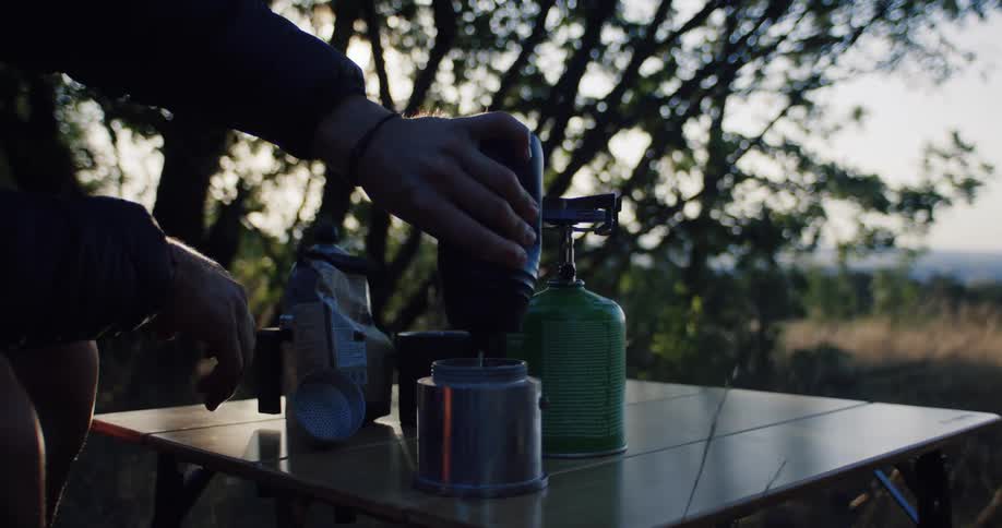 A camper grinds fresh coffee beans with a manual grinder into a moka pot in the early morning sunlight