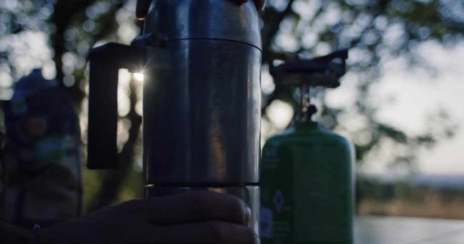A person's hands slowly assemble a metal moka pot on a camping table with the sun flaring through the trees in the background