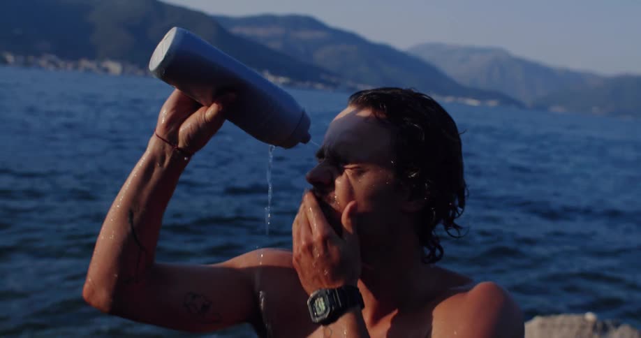 A shirtless man with a mustache cools off by pouring water from a bottle on his face by the sea in slow motion