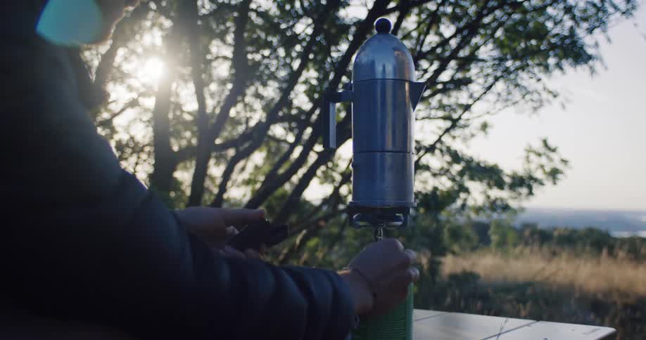 A camper lights a portable stove to brew coffee in a moka pot outdoors as the morning sun shines through the trees