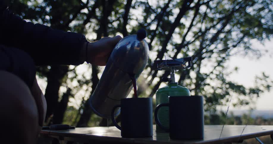 A person pours steaming hot coffee from a moka pot into a mug while camping in the woods at sunrise