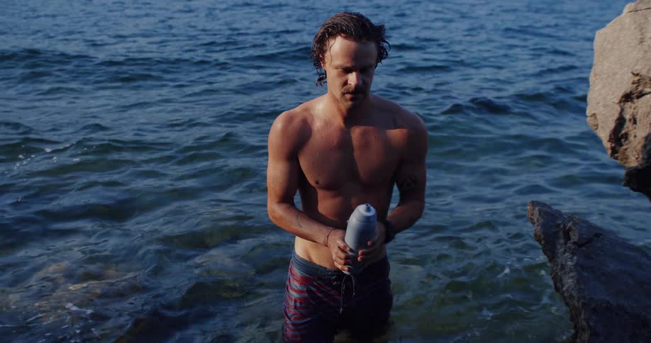 A man in the sea takes a water bottle from a friend and pours it over his head to cool off in the summer sun, captured in slow motion