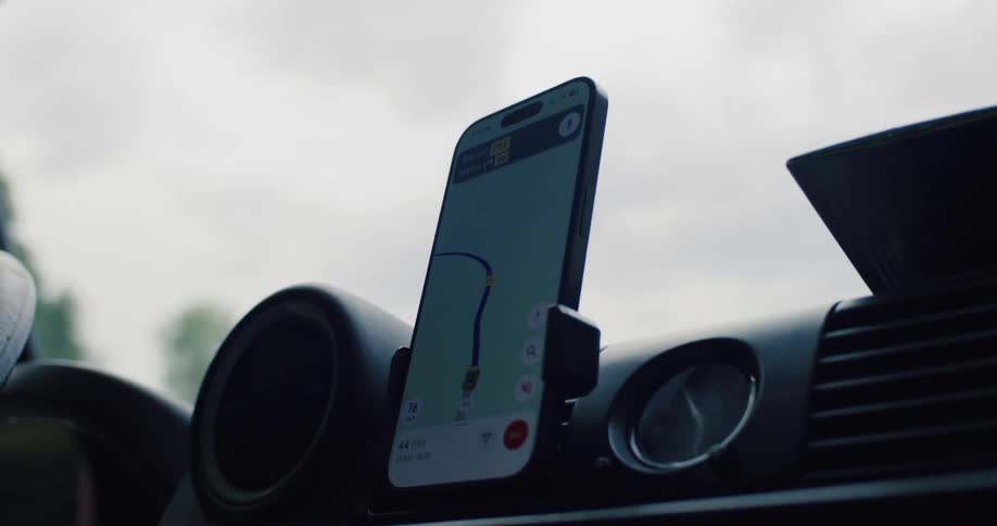 A smartphone with a GPS navigation app guides the way from a car's dashboard during a road trip through a scenic, hilly landscape