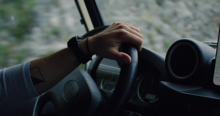 Close-up of a man's tattooed arm and hand steering a vehicle on a scenic road trip through the countryside
